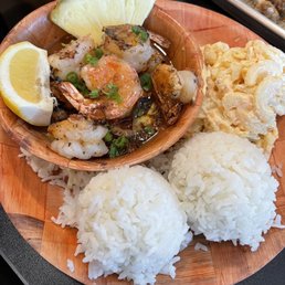 A Hawaiian cuisine plate includes grilled shrimp garnished with chopped green onions, served with white rice, macaroni salad, and fresh pineapple and lemon wedges. The dish is presented on a round wooden platter, with additional food items visible in the background, highlighting traditional Hawaiian dishes with an Asian twist.