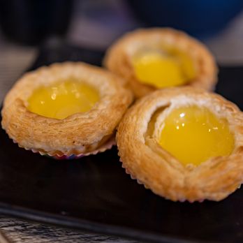 IT DIMSUM - Baked Egg Tart ($5) - Three baked egg tarts with golden, flaky pastry and yellow custard filling are arranged on a black square plate. - Dim Sum Near Me - Albuquerque, New Mexico