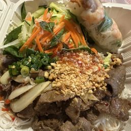Lemongrass Beef Noodle Soup / Bun Bo Hue - The image shows a Vietnamese fast food meal in a takeout container, including sliced beef topped with chopped green onions, crushed peanuts, and sauce, accompanied by shredded lettuce, carrots, and herbs. Two spring rolls with rice noodles and vegetables are also present, all arranged on a bed of rice noodles. The meal emphasizes fresh ingredients and traditional flavors typical of Vietnamese cuisine.