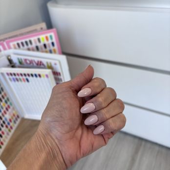 Tiny Nails - Nude polish on acrylics. Done by Tony. - A hand with well-groomed, nude-colored acrylic nails is positioned in the foreground of the image. The background includes color sample displays and a reception desk with the partial word 'RECEP' visible, indicating a nail salon environment. The setting appears clean and organized, with the focus on the manicured nails. - Nail Salons Near Me - Chicago, Illinois