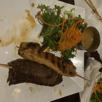 Ditmars Thai - Beef and Chicken Satay - The image shows a white square plate with two skewers of grilled meat, one with a chicken piece and the other with beef. The plate also contains a small side salad with leafy greens and shredded carrots, and a small bowl of dipping sauce. The setting appears to be a dining table with a white tablecloth and some paper napkins nearby. - Thai Near Me - Astoria, New York
