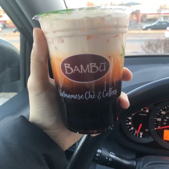 BAMBU - Thai tea with boba - A person is holding a plastic cup of Thai tea with boba from BAMBU, a Vietnamese chè and coffee business, inside a car. The cup features the BAMBU logo and the text 'Vietnamese Chè & Coffees.' The background shows a parking lot and street scene with cars and buildings, indicating the photo was taken outdoors during daylight. - Bubble Tea Near Me - West Allis, Wisconsin