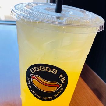 Dogos VIP - Mango lemonade was perfect - A yellow lemonade beverage in a clear plastic cup with a black straw and a sealed lid. The cup has a black circular sticker with a hot dog graphic and the text 'DOGOS VIP' and 'MEAN DOGS. NICE BUNS.' The drink is placed on a wooden table near a window, with natural light illuminating the scene. - Hot Dogs Near Me - Albuquerque, New Mexico