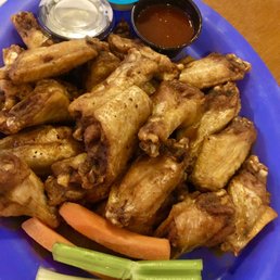 Wings - A blue plastic plate contains a large serving of fried chicken wings, with some celery and carrot sticks at the bottom. Two small containers hold dipping sauces, one with a dark sauce and the other with a creamy, possibly ranch, dressing. The plate is placed on a wooden table, and part of another dish is visible in the top left corner.