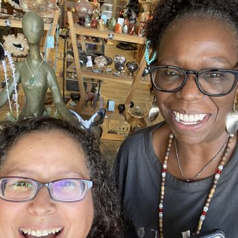Lovestones - Two women wearing glasses are smiling in a shop that sells crystals, fossils, and decorative items, with shelves and displays in the background. - Spiritual Shop Near Me - Menifee, California