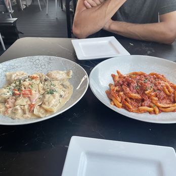 Sogno Wheaton - Lobster Cappellacci and Cavatelli Salsicce - Two pasta dishes are on a black table: one with creamy sauce and seafood, the other with tomato sauce and pasta. A person is partially visible. - Italian Near Me - Wheaton, Illinois