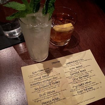 The Eddy - Dew You Mind & High Horse - A tall glass contains a cocktail garnished with fresh mint leaves and a lemon wedge, placed on a dark wooden table. Next to it is a short glass filled with ice and a lemon slice, likely a different drink. A handwritten menu listing various cocktails and their ingredients is also visible, indicating a bar setting. The background includes a glass of water and other bar items. - Cocktail Bars Near Me - Providence, Rhode Island