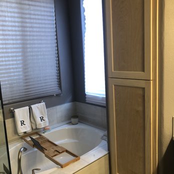 J J 'll Do It - New storage cabinet. - The image shows a bathroom with a built-in bathtub surrounded by beige tiles. A wooden bath tray rests across the tub, holding a black object. Two windows with blinds are positioned above the tub, allowing natural light to enter. To the right, a tall wooden cabinet with a light finish is visible. Two white towels with the letter 'R' hang on a towel bar above the tub. The overall decor includes dark-colored walls and neutral tones. - General Contractors Near Me - Clovis, California