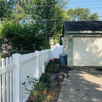 Advanced Fence & Gate - The image shows a backyard area with a white picket fence on the left side, running parallel to a concrete driveway. Several potted plants with green foliage and pink flowers are placed along the fence. A gray trash bin is positioned near the garage, which has a closed white door and a small exterior light. Tall trees with green leaves are visible in the background, and the sky is clear and blue. Shadows from the trees cast on the driveway, indicating sunlight. - Fences & Gates Near Me - Mount Prospect, Illinois