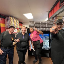 Employees find my phone! - Four employees are inside a pizza restaurant, standing in front of a Pepsi refrigerator and stacked pizza boxes. They are smiling and making gestures towards the camera. The background includes a red and yellow wall, shelves with pizza boxes, and a ceiling with fluorescent lighting.