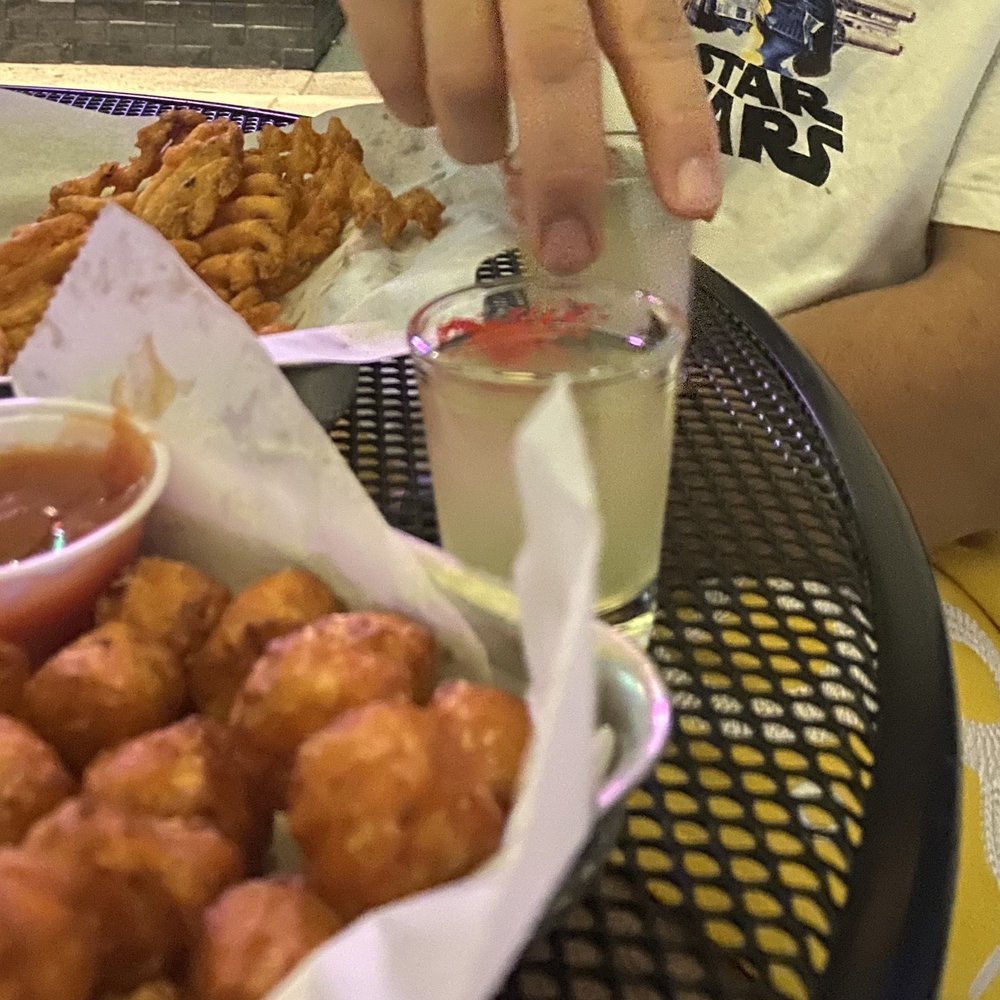 The Avenue - A man grabbing for his yellow shot with waffle fries and sweet potato tots on table - Burgers Near Me - Boston, Massachusetts The Avenue - A man grabbing for his yellow shot with waffle fries and sweet potato tots on table - Burgers Near Me - Boston, Massachusetts