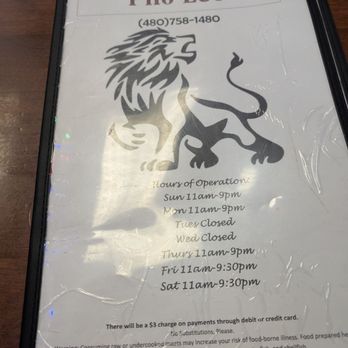 Pho Leo - Decent menu, there aren't so many choices that you feel overwhelmed. - The image features a menu cover for Pho Leo, a Vietnamese restaurant, displaying the restaurant's name, phone number, and hours of operation. The menu has a lion and dragon logo printed in black, with the hours listed for each day, including closures on Tuesday and Wednesday. The menu is encased in a black frame with a plastic cover that has some wrinkles and reflections. Additional text notes a surcharge for card payments and food allergy warnings. - Vietnamese Near Me - Mesa, Arizona