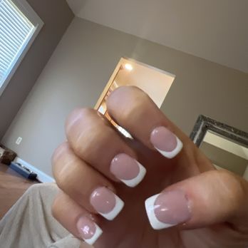 Amazing Nails - The image features a hand with neatly manicured nails displaying a French manicure, characterized by pink nail beds and white tips. The hand is positioned in front of a room with beige and gray walls, a window with blinds, and a mirror. The room has a ceiling light and wooden flooring, with some objects visible on the floor near the wall. - Nail Salons Near Me - Pelham, Alabama