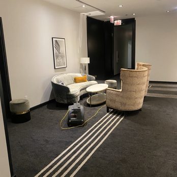 Pendry Chicago - The image shows a hotel lobby seating arrangement with a velvet sofa, two beige upholstered armchairs, a small oval coffee table, and a floor lamp. The area is decorated with a black and white framed photograph on the wall. The floor is carpeted with a dark pattern, and there is a cleaning machine on the carpet. The background includes a black door and an exit sign, with ceiling lights illuminating the space. - Hotels Near Me - Chicago, Illinois