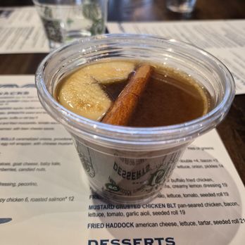 Cisco Brewers - Hot cider - A clear plastic cup contains hot cider, garnished with a cinnamon stick and a slice of apple. The cup is placed on a restaurant menu, with other drinks and menus visible in the background. The setting appears to be a dining area at a restaurant or cafe. - Brewpubs Near Me - Boston, Massachusetts