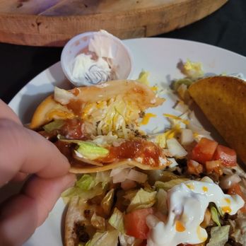 The Rusty Nail Saloon - Tacos - A white plate holds two tacos filled with ground beef, lettuce, tomatoes, shredded cheese, and sour cream. A small cup of sour cream is also on the plate. A side of tortilla chips is partially visible on the right side of the plate. The plate is on a table with a wooden surface in the background, and a glass of dark soda is visible on a wooden tray. - Pizza Near Me - Lostant, Illinois