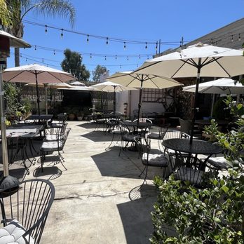 Cafe BT - The image shows an outdoor patio at Cafe BT in Walnut, CA, with multiple black metal tables and chairs, some with cushions. Large umbrellas provide shade over the tables, and string lights are strung across the area. There are plants and trees along the perimeter, creating a garden-like atmosphere. The concrete floor has visible cracks, and a patio heater is present on the left side. The setting appears suitable for dining or socializing during daytime. - Coffee & Tea Near Me - Walnut, California