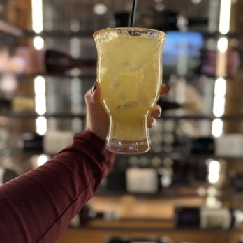ChopHouse Grille - A person wearing a long-sleeve shirt holds a tall, curved glass filled with a yellow cocktail and ice cubes. The glass has a salted rim and a black straw. The background is a blurred bar interior with shelves and bottles, emphasizing the drink as the focal point. - New American Near Me - Wakefield, Rhode Island