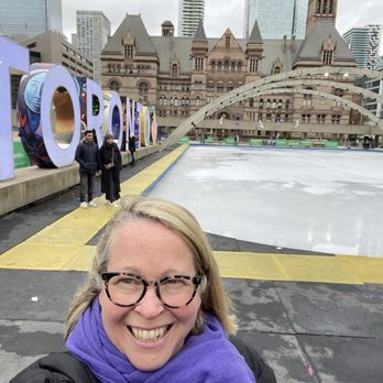 Nathan Phillips Square - A woman with glasses and a purple scarf is smiling and taking a selfie at Nathan Phillips Square in Toronto. Behind her, there is an outdoor ice skating rink, with a large, colorful 'TORONTO' sign and a historic building, Toronto City Hall, with a distinctive architectural style. The area is surrounded by tall modern buildings, and the sky appears overcast. - Landmarks & Historical Buildings Near Me - Toronto, Ontario
