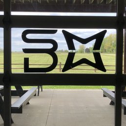 A window with a logo consisting of the letters 'SM' and a star shape, viewed from inside a pavilion or shelter. The background shows a grassy field, a wooden fence, and trees under a partly cloudy sky. The window is framed by structural elements of the pavilion, including a wooden roof and benches outside.