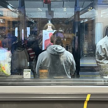 RTSP - Firing range - A person in a gray hoodie with headphones is at a shooting range, viewed through a glass window with reflections and other people visible. - Gun/Rifle Ranges Near Me - Randolph, New Jersey
