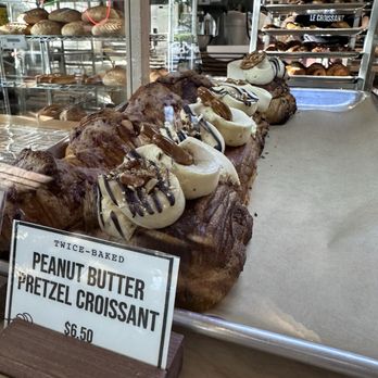 Le Croissant - Peanut Butter Pretzel Croissant - A tray of peanut butter pretzel croissants topped with chocolate drizzle and nuts, displayed in a bakery case at Le Croissant in Logan, UT. - Bakeries Near Me - Logan, Utah