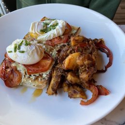 Poached Eggs - A white plate holds poached eggs garnished with chopped chives, placed on tomato-topped toast. Next to the eggs, there are fried potatoes with red peppers. The dish is presented on a marble table, with a person wearing a green jacket visible in the background.