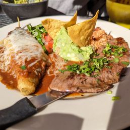 Chile Relleno - A white plate holds carne asada garnished with chopped herbs, a chile relleno covered in sauce and melted cheese, a serving of rice, fresh guacamole, and two tortilla chips. The dish is set on a dark outdoor table with bowls and other items partially visible in the background.
