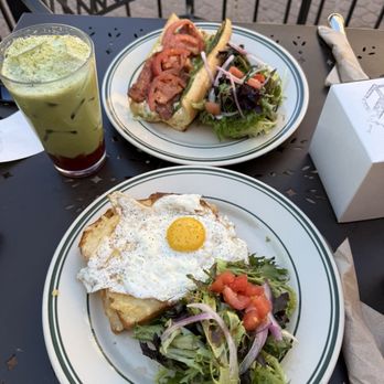 Maisonette - Croquet madame and the Roma. Strawberry matcha latte - The image shows an outdoor dining setup with two plates of food and a drink on a black metal table. One plate contains a fried egg with a salad of mixed greens, tomatoes, and onions. The other plate features a sandwich with bacon, sliced vegetables, and a side salad. A tall glass with a layered green drink, possibly a matcha or vegetable smoothie, is also present. The setting includes patio furniture and a paved ground. - Bakeries Near Me - Boulder, Colorado