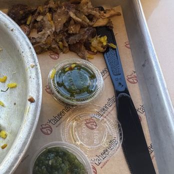 Taboonette - That's what I took off my plate I can't eat that despite of this very small amount of meat - The image shows a metal tray with leftover cooked meat, possibly from a Mediterranean dish, on a paper placemat branded with Taboonette. Three small plastic containers of sauces are placed on the tray: one with green sauce, and two with brownish sauces. A black-handled knife rests on the tray, and part of a metal plate with some remaining food is visible on the left side. The setting appears to be a casual dining environment. - Mediterranean Near Me - Brooklyn, New York