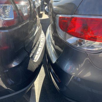 Cactus Jack's Auto - The image shows two cars parked side by side in a parking lot, with their rear ends very close, almost touching. The vehicle on the left is black, and the one on the right is gray. The parking lot has multiple other cars visible in the background, and the scene is illuminated by sunlight, creating reflections on the car surfaces. - Car Dealers Near Me - Glendale, Arizona