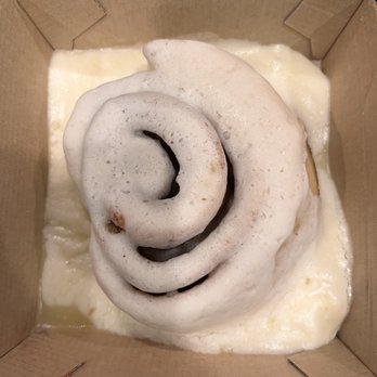 Cinnaholic - Old Skool Roll on last visit quite smaller. - A cinnamon roll with white icing is positioned inside a cardboard takeout box. The roll is spiral-shaped, with visible layers of dough and cinnamon filling. The icing appears to be cream cheese frosting, covering the top of the roll and spreading slightly onto the box. The box is placed on a dark, speckled countertop surface. - Bakeries Near Me - Gambrills, Maryland