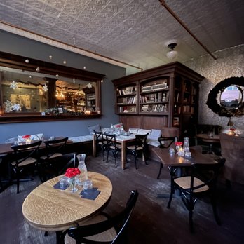 205 Dry - The image shows a restaurant interior with dark wooden furniture, including tables and chairs, arranged in a cozy setting. A large mirror with snowflake decorations is mounted on the wall, reflecting warm lighting from inside the restaurant. There is a wooden bookshelf filled with books and a decorative mirror with a wreath-like frame on the wall. The ceiling has a patterned texture, and the overall ambiance suggests a warm, inviting atmosphere suitable for casual dining or gatherings. - Speakeasies Near Me - Binghamton, New York