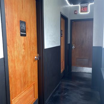 SubKO - View of bathrooms -  clean space - A hallway with three doors, two of which are restrooms with wood finish doors and black frames. The doors have restroom signs, and there is an illuminated exit sign above the hallway. The walls are painted in a light color with a black lower section, and the floor has a dark, marbled appearance. - Sandwiches Near Me - Lisle, Illinois