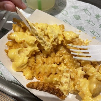 Shake Shack Southdale - Cheese Fries - Crinkle-cut French fries topped with melted cheese sauce are presented on a paper tray, with a plastic fork and a wooden utensil. A drink with a green straw is visible in the background. The setting appears to be a casual dining environment, likely at Shake Shack in Minneapolis, MN. - Fast Food Near Me - Minneapolis, Minnesota
