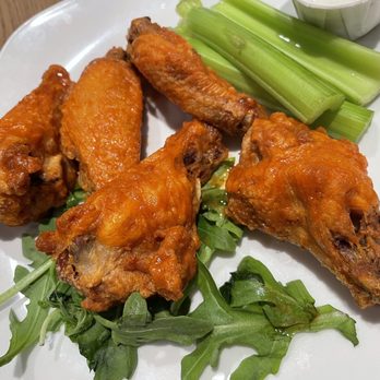 Maxi's Restaurant - Buffalo wings - A white plate holds several buffalo chicken wings coated in orange buffalo sauce, arranged with some on a bed of greens. There are celery sticks and a small container of dipping sauce in the corner. The dish appears to be served in a restaurant setting. - New American Near Me - Modesto, California