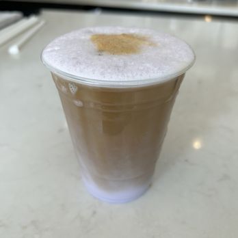 Concept Coffee - Iced Ube latte - A clear plastic cup holds an iced ube latte with a layer of foam and a sprinkle of cinnamon or similar spice on top. The drink is placed on a light-colored, smooth countertop, with a blurred background showing various coffee shop items and equipment. The beverage appears to be a specialty Asian-inspired latte with a purple hue visible through the cup. - Coffee & Tea Near Me - Queens, New York
