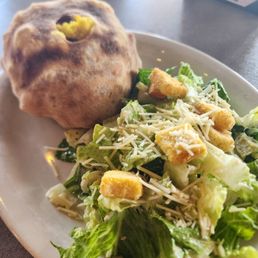 Brunch burger and Caesar salad - A Caesar salad with croutons and shredded cheese is served on a white plate, accompanied by a bread roll with a small opening on top.