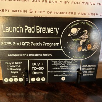 Launch Pad Brewery - Reward program! - The card is black with white text and a space-themed graphic of an astronaut on a tractor, wearing a helmet and suit, surrounded by planets and stars. It details a 2025 second quarter patch program, including missions such as buying beers and taking photos at the brewery. The card provides contact information, social media instructions, and a list of beer tap numbers for a rewards program. - Breweries Near Me - Aurora, Colorado