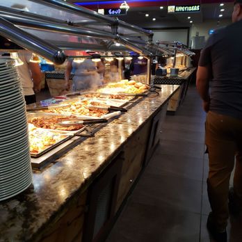 King Buffet - Rows and rows of fried foods - A buffet setup in a restaurant features a long granite counter with heated trays containing various cooked dishes. Steam rises from some trays, indicating hot food. Plates are stacked on the side for diners to serve themselves. Several customers and staff are visible in the background, with signs overhead indicating sections like soup and appetizers. The environment has a modern interior with colorful lighting and a busy atmosphere. - Buffets Near Me - Plano, Texas