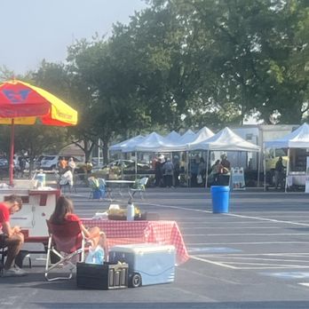 Yukich Fields - Farmer's Market - A farmer's market is set up in a parking lot with several white tents serving as vendor booths. People are walking around and browsing the stalls, while some sit near a red and yellow umbrella providing shade. The scene includes trees in the background and parked cars, indicating an outdoor community event. - Parks Near Me - Evergreen Park, Illinois