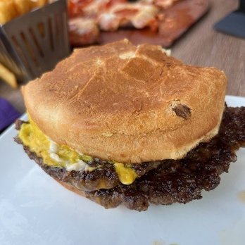 Ithaca Ale House - Smash Burger - A breakfast sandwich with a golden-brown biscuit, scrambled eggs, and a sausage patty is placed on a white plate. The biscuit appears soft with a slightly crispy exterior. The background shows other food items, including slices of meat and fries, on a wooden table. The image captures a close-up view of the sandwich, emphasizing its ingredients and texture. - New American Near Me - Ithaca, New York