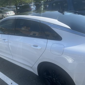 Tint Plus - Tint Driver side - A white sedan with black wheels and tinted windows is parked in a marked parking space in a parking lot. The vehicle's side profile is visible, with the door handles and fuel cap clearly shown. The parking lot contains multiple other cars, including SUVs and sedans, and is bordered by trees. The image captures a sunny day, with reflections of nearby buildings and trees visible on the car's surface. - Home Window Tinting Near Me - Woodbridge, Virginia