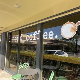outside of a coffee shop - A coffee shop storefront window features the phrase "i need coffee" and coffee cup decals, with outdoor seating visible.