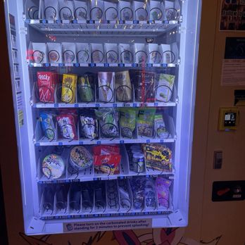 Yamada Vending - The vending machine features a glass front displaying various snacks, drinks, and Pokémon-themed items. The top row has small Pokémon collectible items, while the middle rows contain snack packages such as Pocky and other snacks. The bottom section includes beverage cans and additional snack items. The machine has a keypad and payment system on the right side, with a sign instructing customers to turn on carbonated drinks after standing for two minutes. The background includes part of a Pokémon-themed design at the base of the machine. - International Grocery Near Me - Eugene, Oregon