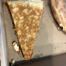 Not Lucky's pizza - A single triangular slice of cheese pizza with a golden-brown crust and melted cheese is placed on a cardboard pizza box. The slice appears to be partially eaten, with some crumbs and small pieces of cheese visible around it. The pizza box surface is plain and slightly textured, with a few crumbs and bits of cheese nearby.