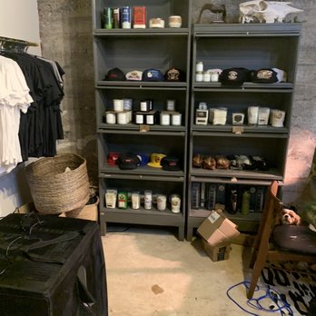 Hide & Hunter - Two black shelving units contain hats, candles, and small items, with a dog peeking from a chair on the right, in a room with a concrete wall. - Leather Goods Near Me - Birmingham, Alabama
