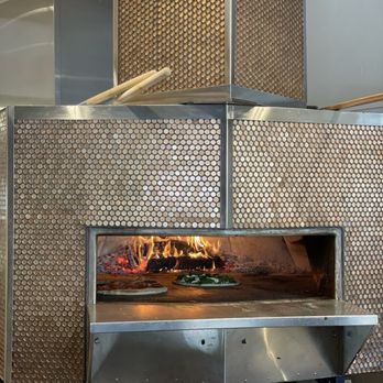 ZA - A stainless steel wood-fired pizza oven features a glass opening revealing two pizzas cooking inside, with flames visible in the background. The oven has a metallic, textured exterior and is situated in a kitchen or pizzeria setting, with some utensils and equipment nearby. - Pizza Near Me - Elm Grove, Wisconsin