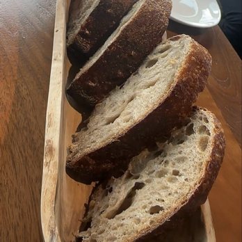 Canoe - Bread - A wooden tray holds several slices of rustic, crusty bread with an airy interior, placed on a table with a white plate nearby. - Canadian (New) Near Me - Toronto, Ontario