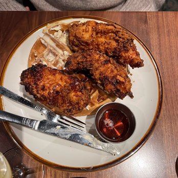 Herbie's Restaurant - A plate with three fried chicken pieces, waffles, and syrup is on a wooden table, with a fork and knife placed on the plate. - New American Near Me - Clayton, Missouri